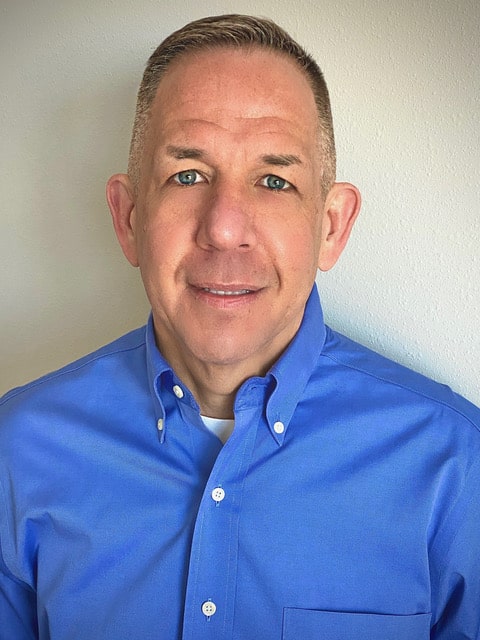 headshot of a man smiling in a blue button down shirt against a beige wall