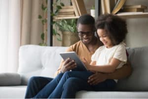 father holds on lap daughter sitting resting on sofa in living room using digital tablet holding mobile computer